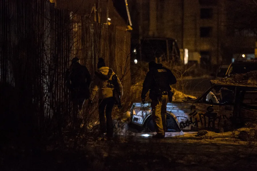 A group of three people walk together in the dark. One shines a flashlight on a car covered in graffiti that doesn't have any tires.