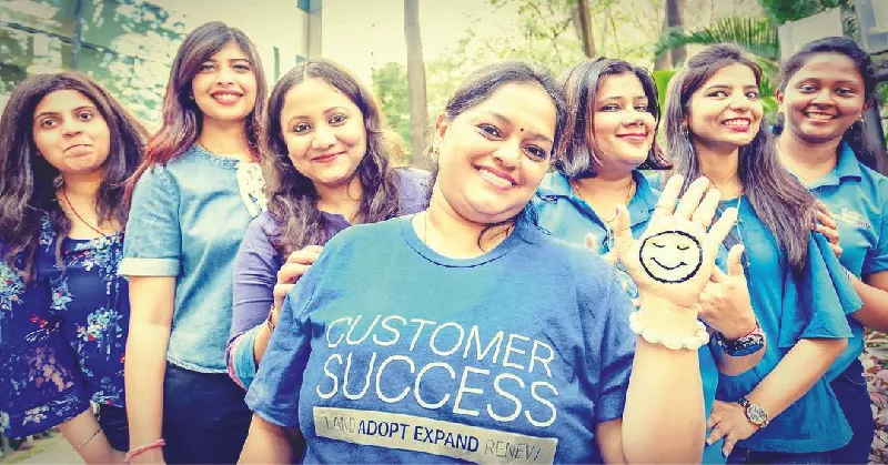 The image shows seven women smiling. The one in the center is wearing a shirt that says “Customer Success.”