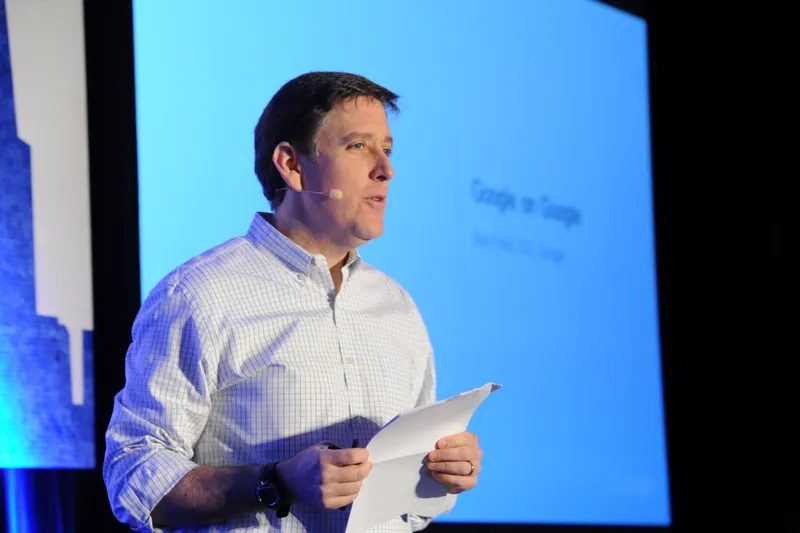 A photo shows Ben Fried speaking during a conference with papers held in his hand.