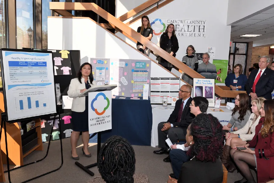 A speaker at a podium next to an easel addresses a roomful of people. A chart with a bar graph and other written information sits on the easel.