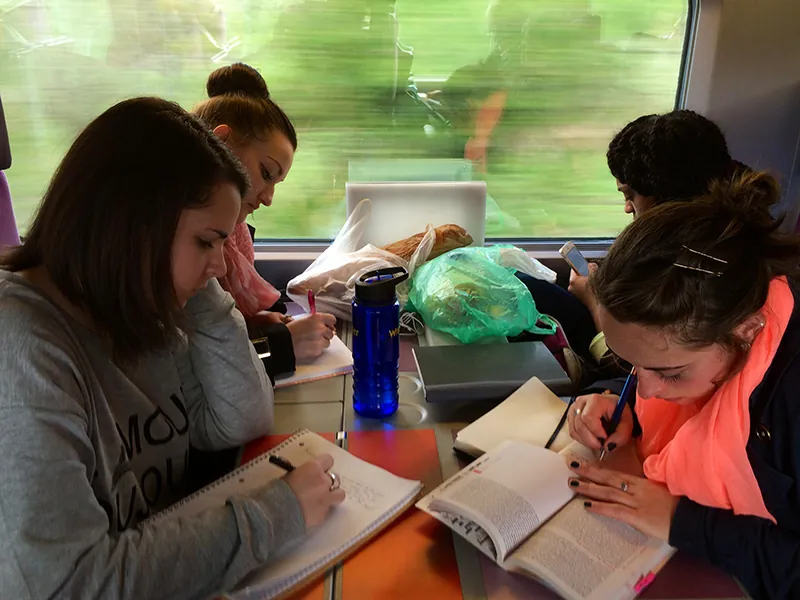 Four people sit at a table on a train. They are reading and taking notes.