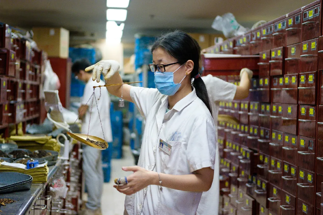 A pharmacist weighs dried plants on a scale while standing in front of a wall of small, square, wooden drawers. Other pharmacists work in the background.