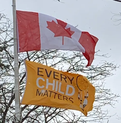 Photo of flag pole with Canadian flag at the top and under it, an orange flag that says Every Child Matters.