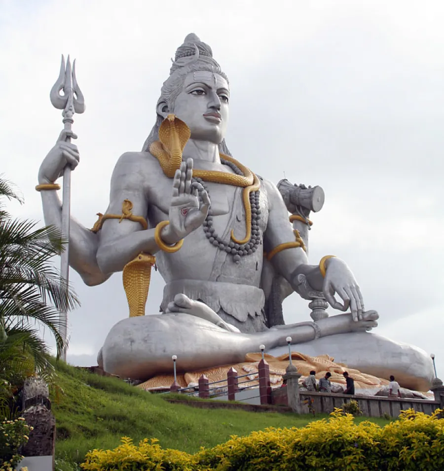 Photo of a stone Shiva with four arms and a gold snake twisting around his neck and one sitting at his side. Shiva is holding a trident and wearing gold bracelets.