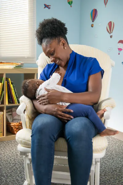 An infant lays across his mother's lap to breastfeed. The mother is sitting in a rocking chair and has her blouse partially unbuttoned.