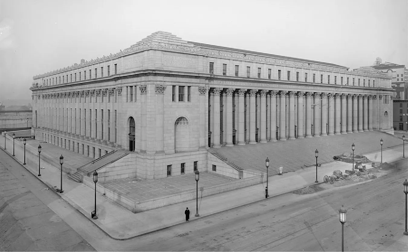 A black and white picture of the corner of a large stone building is shown. Tall columns decorated at the top line the outside of the building on the two sides shown. Rectangular windows line the top of the building and the corner is bricked with an inscription over the colonnade on both sides of the corner. An arched doorway is shown on the left side with stairs leading up to it from the sidewalk. A recessed arched alcove is seen on the other side of the corner. Stairs run the length of the building on the right side. Lampposts line the street on both sides and a road is seen in the forefront on both sides of the building. Other buildings can be seen in the background and a lone figure stands at the corner in a long black coat and hat. Some wheels and piles of dirt can be seen on the right side of the building in front of the stairs.