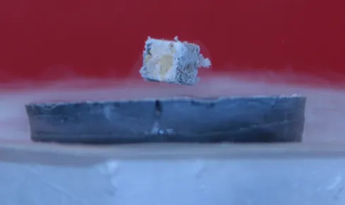La imagen muestra una fotografía de un pequeño imán en forma de cubo que levita sobre la superficie de un superconductor.