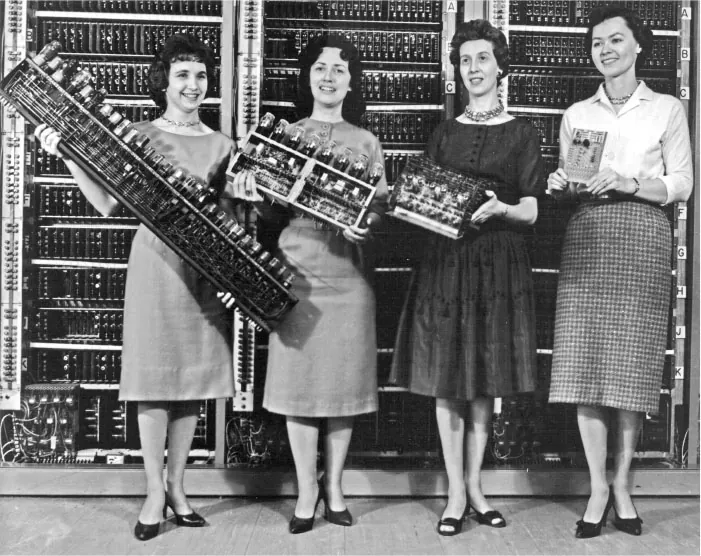 This black-and-white image shows four women from the early computing era standing in front of a large computer system. Each woman is holding different pieces of hardware, such as circuit boards, vacuum tubes, and other computer components.