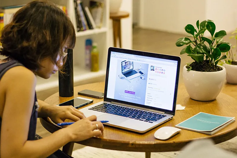A student sits at a table in front of a laptop computer. The student holds a pen in their right hand. A plant, computer mouse, and notebook rest on the table. 