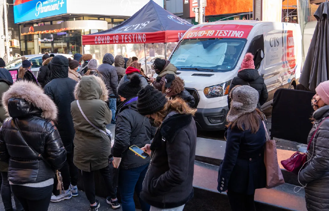 A long line of people wearing winter coats and hats stand outside in the street near a van and tent labeled Covid 19 Testing.