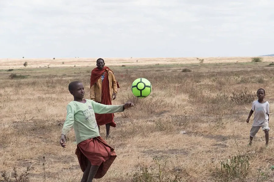 Image of two children and an adult playing with a neon green soccer ball in a desert area.