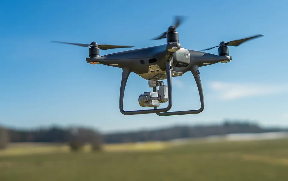 A black quadcopter drone with a camera hovers in mid-air over a blurred field and trees under a clear blue sky.