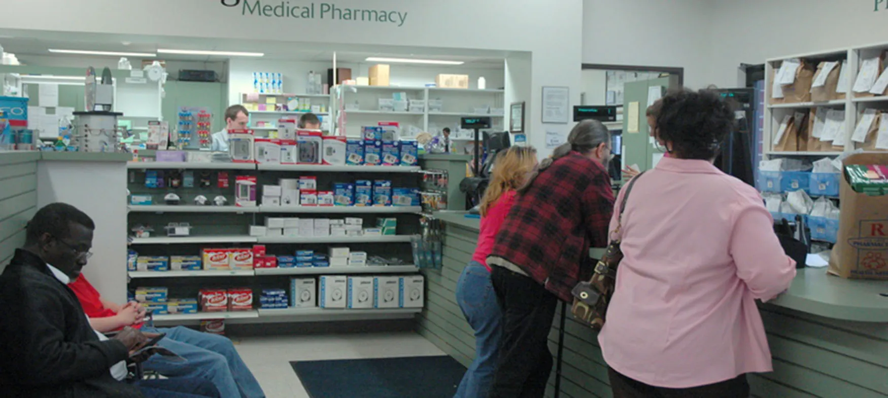 Photo of individuals standing at pharmacy counter while others wait in chairs.