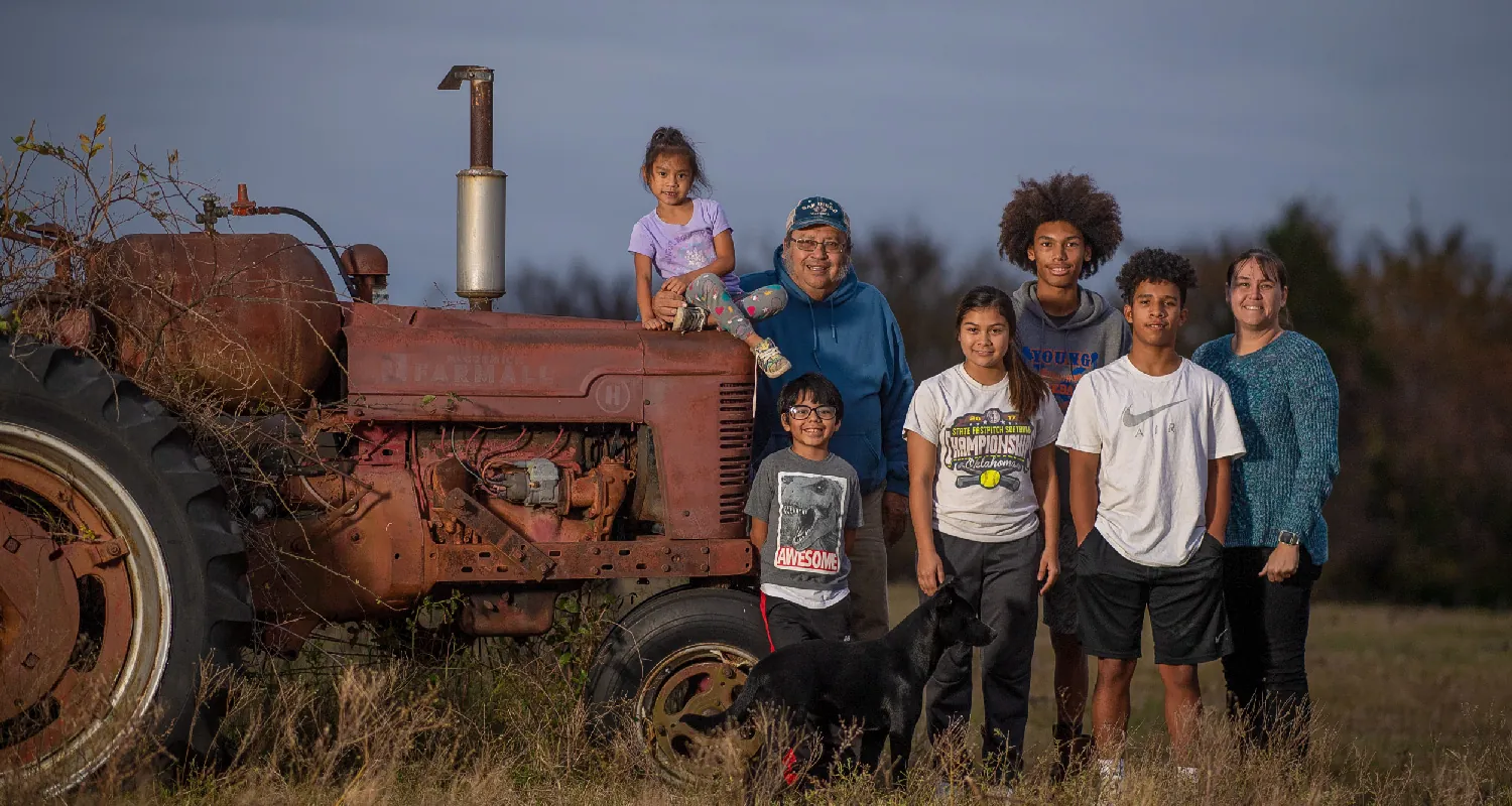Seven people, ranging in age from from young childhood through middle age, stand outside near a rusted tractor.