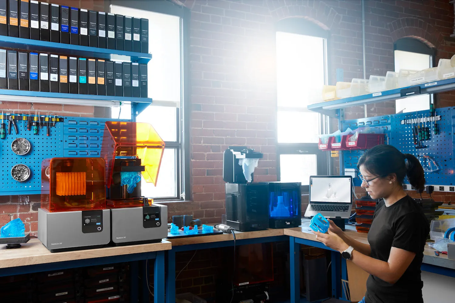 A person holds and looks at a part in a fabrication lab containing multiple 3D printers, a computer, and other equipment.