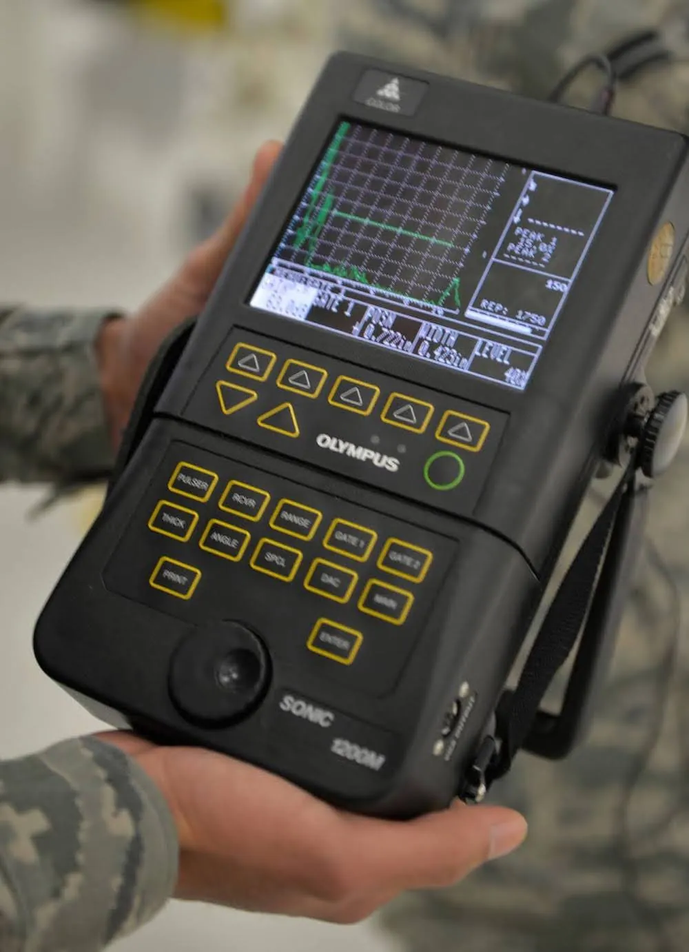 A person holds a device that is about a foot long and six inches wide, with several buttons and controls and a screen displaying a grid display and some data.