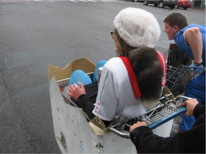 A young individual in a shopping cart being pushed in a parking lot.