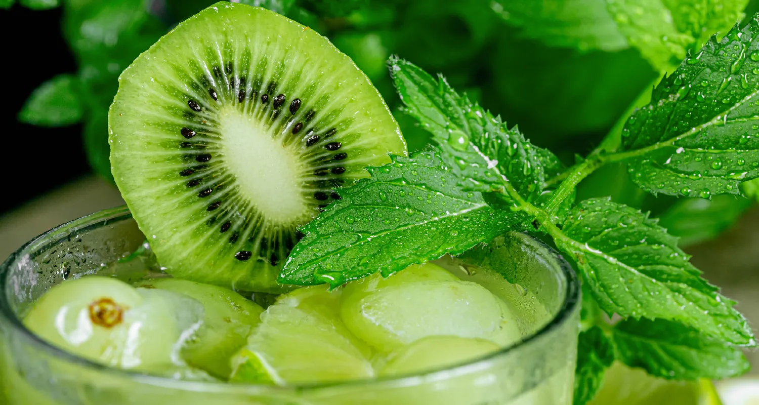 Close-up of the top of a glass filled with a liquid, green fruit, including grapes and kiwi, and a mint leaf.