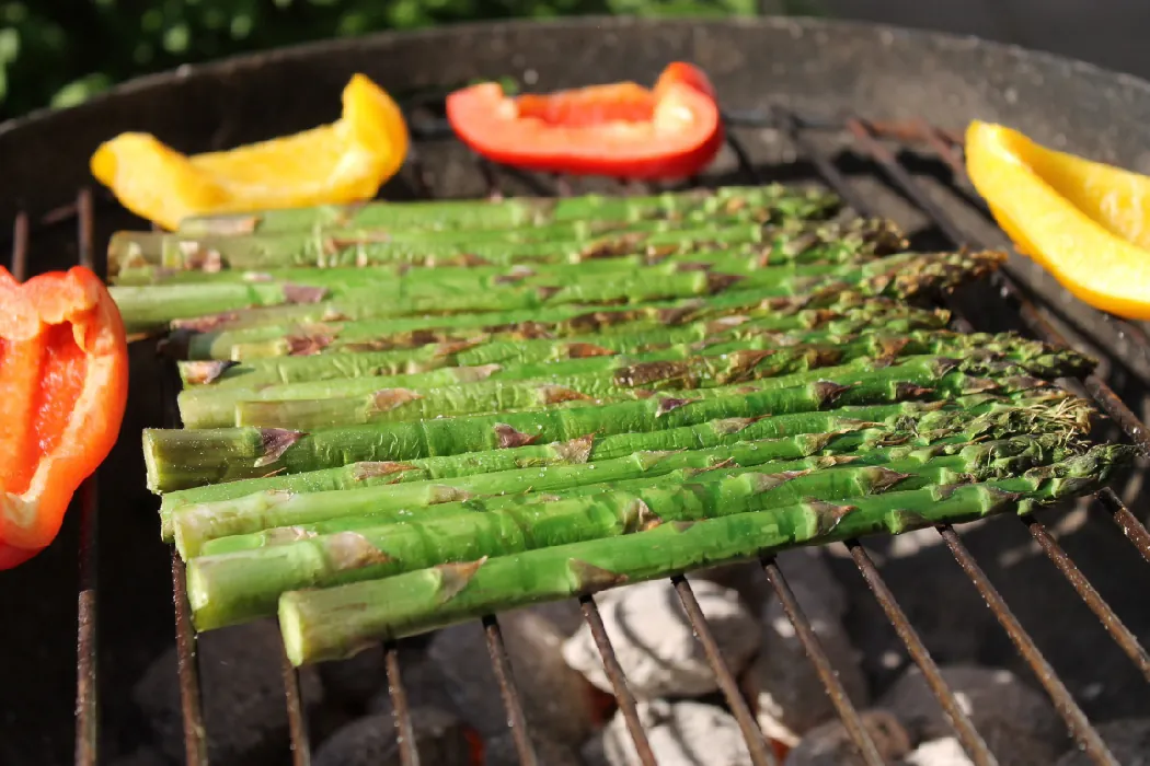 Fresh asparagus spears and quartered red and yellow peppers on a charcoal grill.