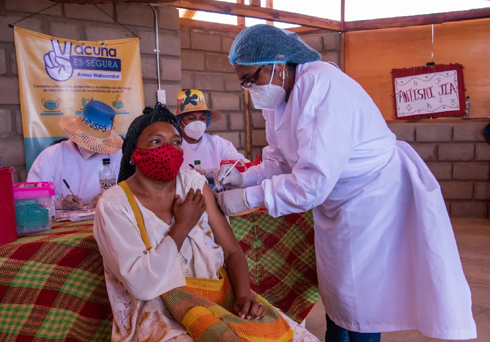 A seated client holds up their sleeve so that a healthcare worker can administer a vaccination. Two other healthcare workers sit at a table behind them. All four people in the photo wear face masks to cover their nose and mouth.