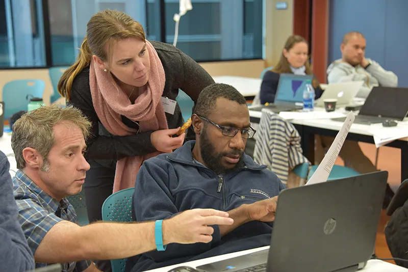 Three people, two seated and one standing, look at an open laptop.