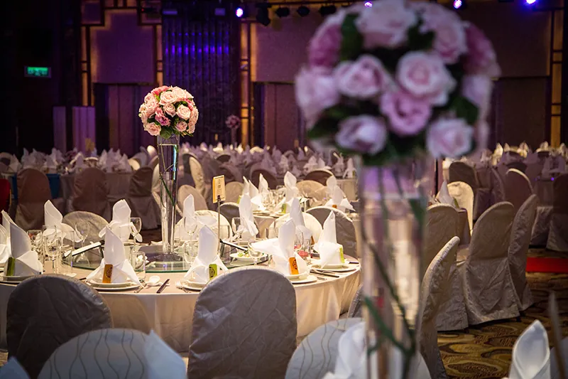 A fancy round table is shown in a banquet room; other similar tables are visible in the background. The table is covered in a table cloth and is set with formal dishes and silverware. Cloth napkins are folded so they stand straight up at the center of the table. A round bouquet of flowers is at the center of the table in a tall narrow vase.