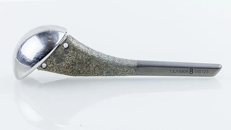At one end of a metal implant is a substantial half sphere, polished smooth. It is attached to a much narrower part that tapers over its length. The tapered part has a rough texture at the top, closest to the ball, and a smoother surface at the narrower end. The implant has a series of small numbers on it and a larger number 8.
