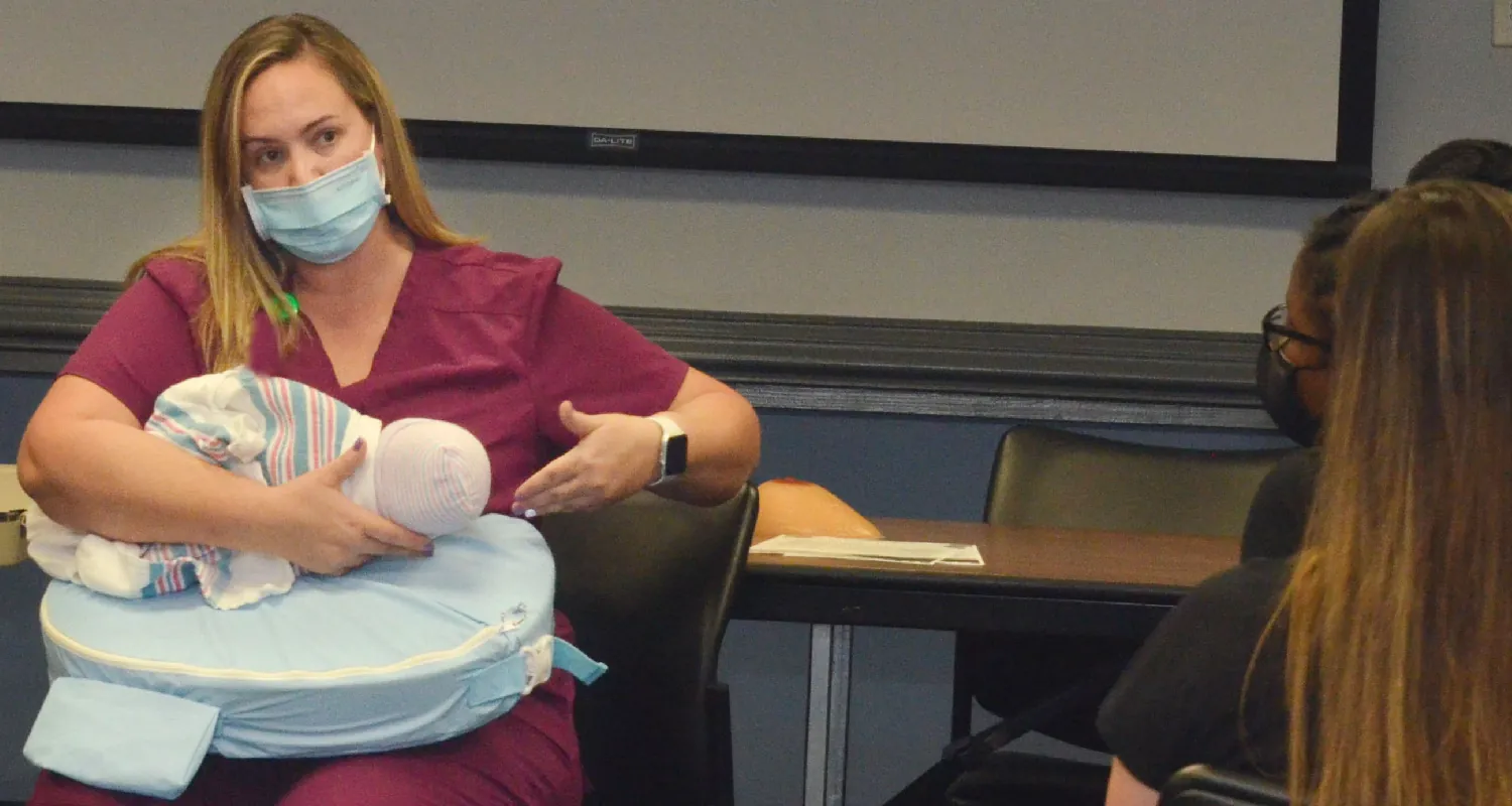 A nurse sits on a chair with a nursing pillow on her lap, holding a baby doll to her chest with one arm, and gesturing toward the doll with the other, while several students observe.