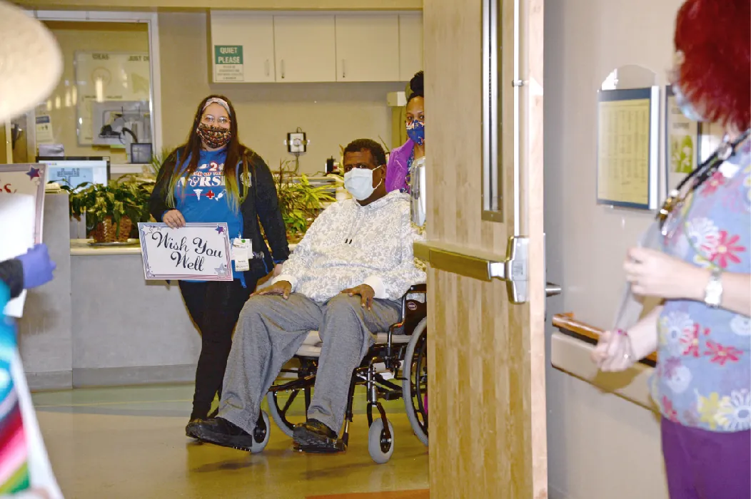 A nurse pushes a client in a wheelchair through a set of doors. Another nurse walks next to them, holding a sign that says Wish You Well.