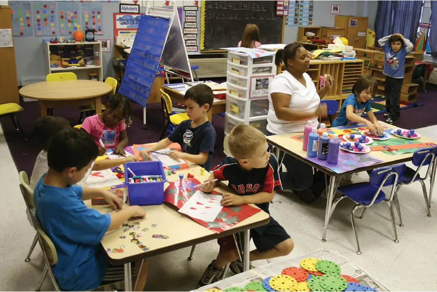 Image of a primary classroom with children and teacher sitting at short, rectangle tables, working on puzzles, coloring, and writing. Other children are in various parts of classroom walking around.
