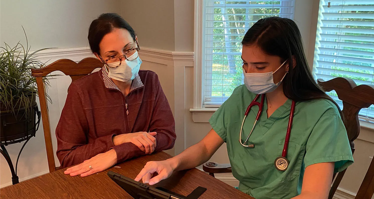 A color photograph of a nurse and patient sitting at a kitchen table, both wearing masks. The nurse is using a tablet while the patient looks on.