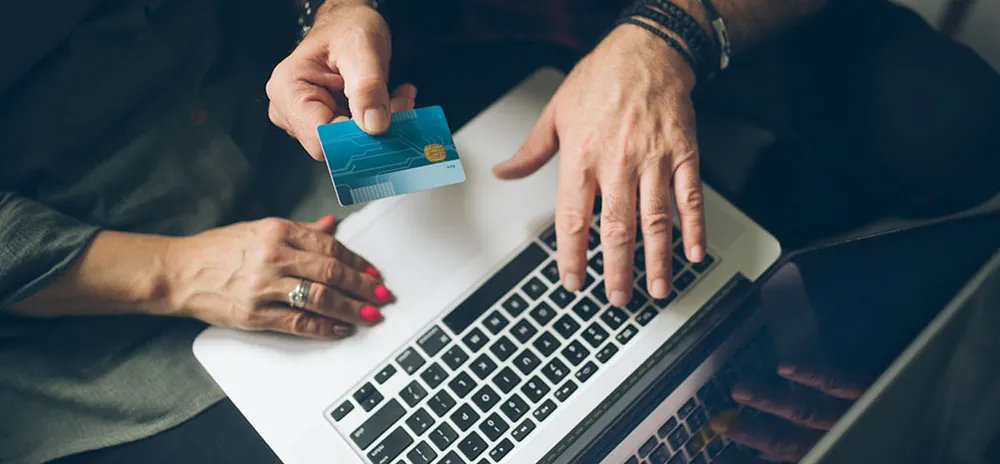 Three hands are shown. Two are working on a single laptop while a third holds a credit card.