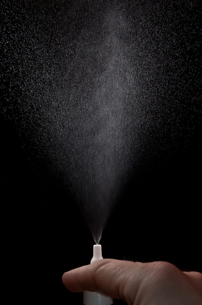 A hand sprays a mist of nasal spray droplets on a black background.