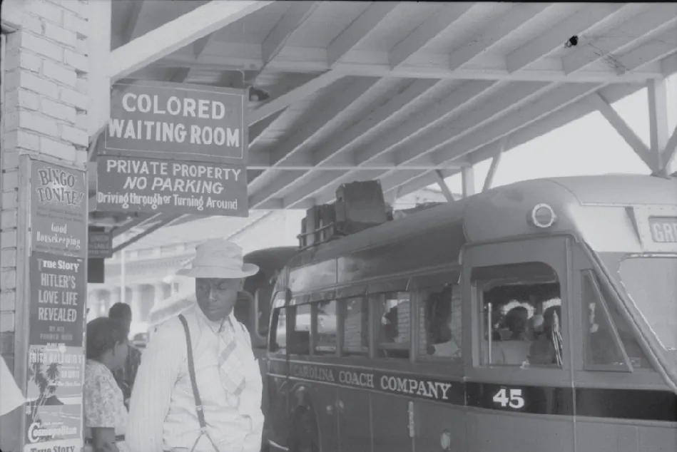 A Black man stands outside next to a parked bus. A sign above him says Colored Waiting Room.