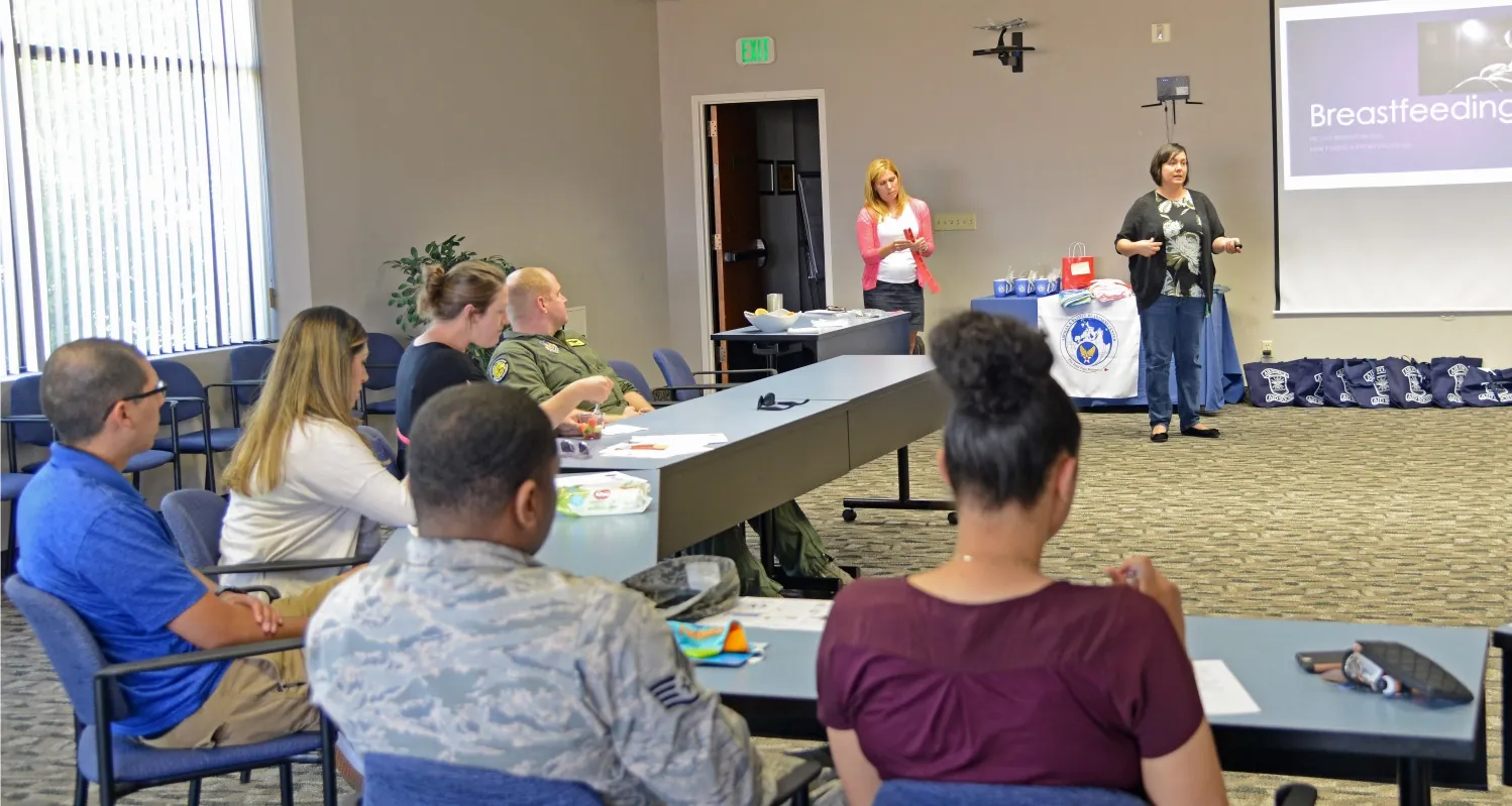 A group of expectant parents sit around tables during a childbirth education class. An instructor stands in the front of the room.