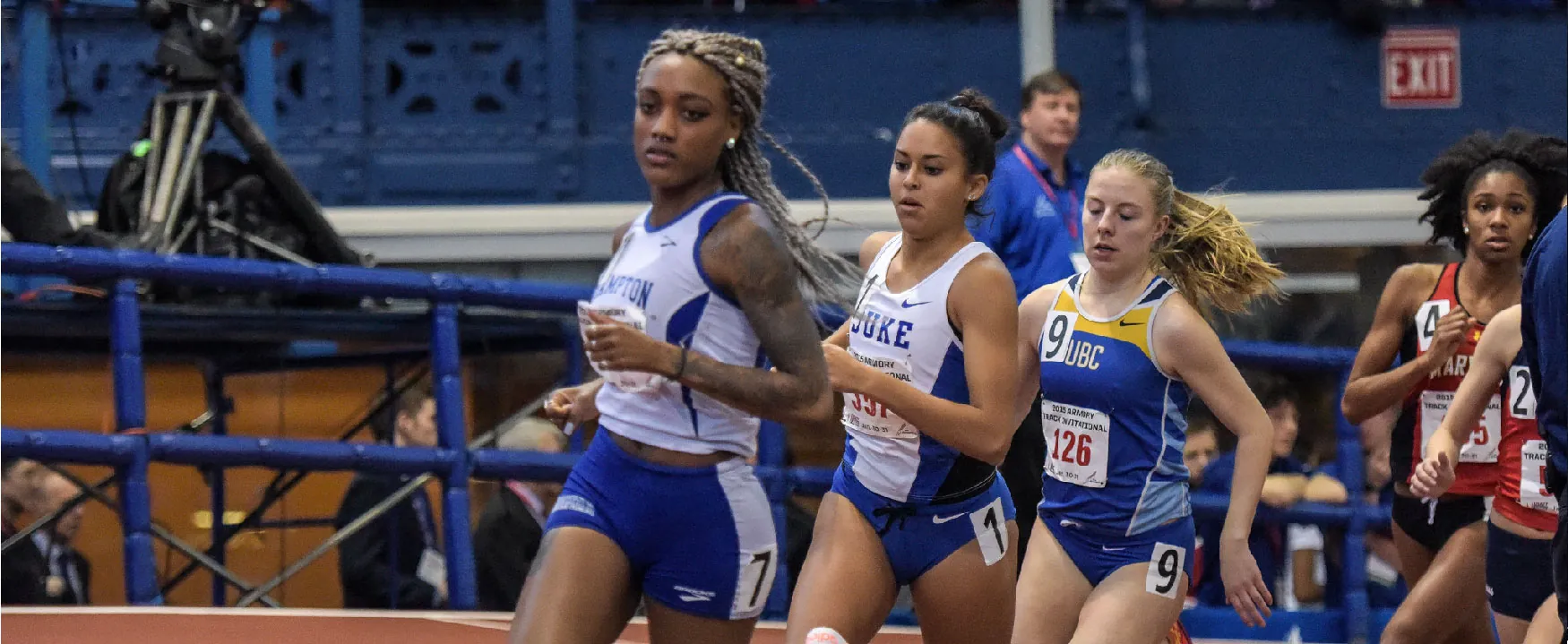 Female athletes from different teams, including Duke and UBC, intensely compete on an indoor track. Runners in blue, white, and red uniforms are captured mid-race, showcasing their focus and speed.