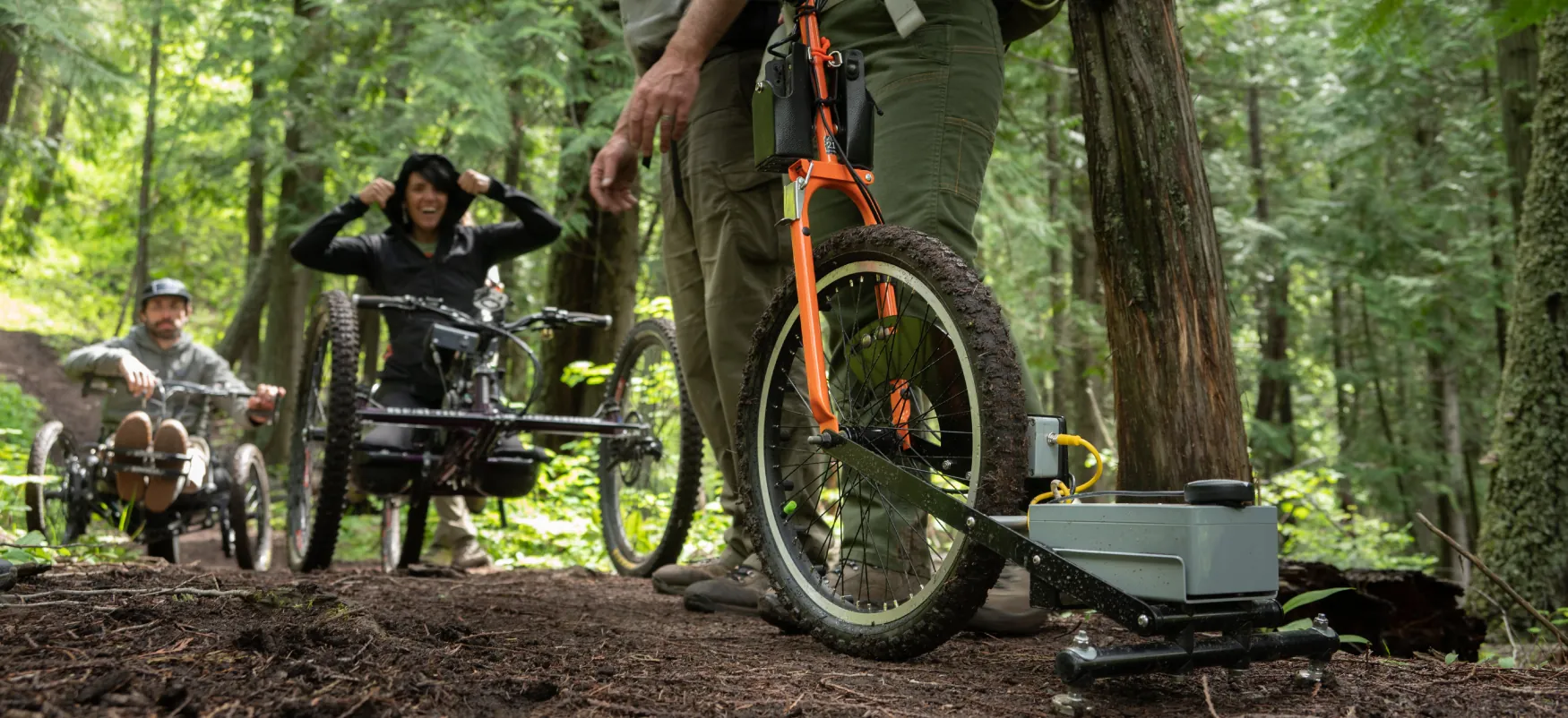A trail in a forest with two individuals seated on three-wheeled mountain bikes using hand cycles. Two more individuals stand by a two-wheeled bike, with a measuring device attached to the wheel.