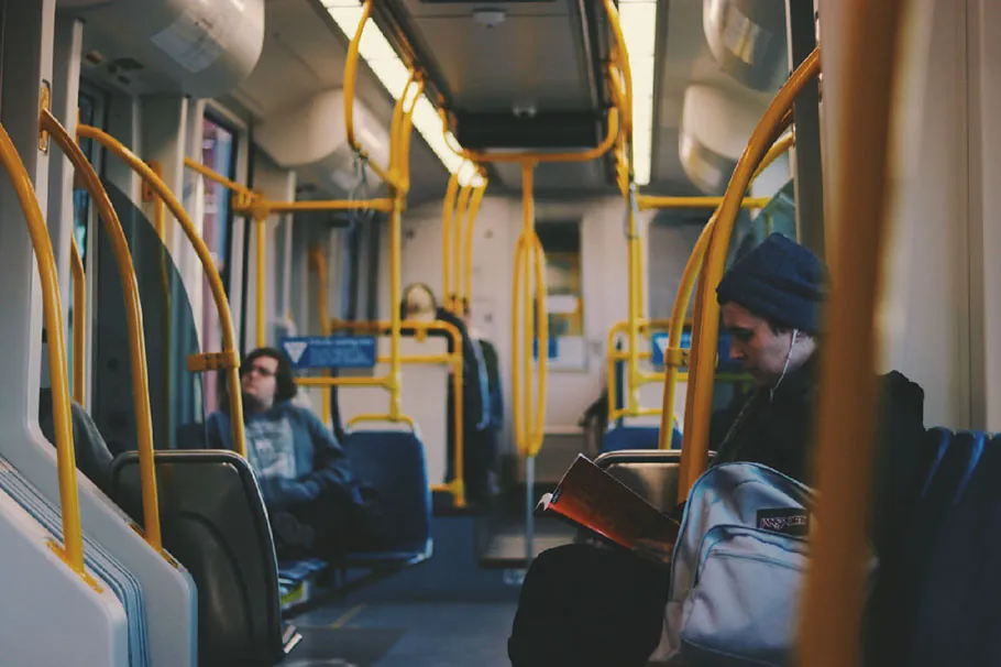 A person with earphones plugged in his ears sits in a train reading a book. Other commuters are seated in the background.