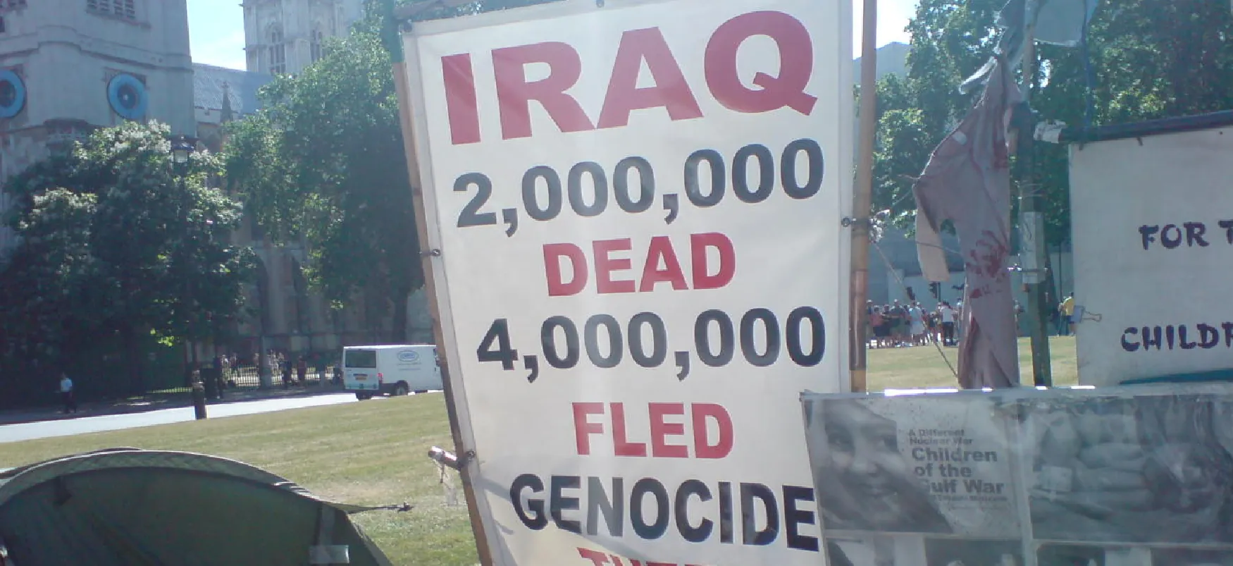 An unseen person holds a protest sign that reads “Iraq,” “2,000,000 dead,” and “4,000,000 fled genocide.”
