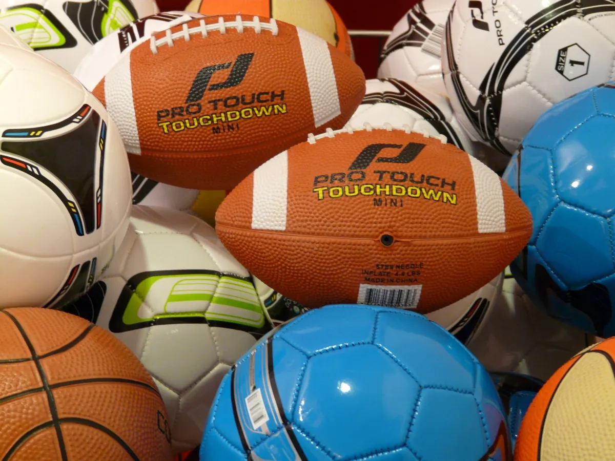 A close-up of various sports balls, including footballs, soccer balls, and a basketball, piled together. The central football has Pro Touch Touchdown printed on it.