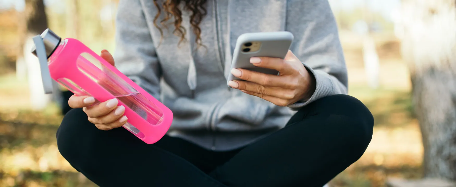 A person sits in a wooded area holding a mobile phone and a water bottle.