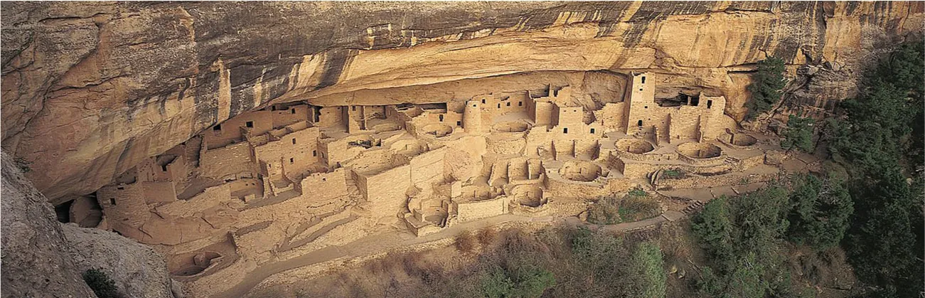 A photo is shown of a black and beige cliff at the top. Below the cliff are seen remains of stone buildings. The buildings run the length of the photo and are stacked in tiers of varying heights, They go deep into the canyon below the cliffs. Windows, and doors can be seen on the buildings, and walls are seen in varying sizes all over. Ten round areas set into the ground are seen toward the right of the canyon. A path is seen running the length of the photo along the front of the structures. Trees are seen at the right of the photo while bushes are seen along the bottom right where the ground dips down. At the left more canyon walls are seen.