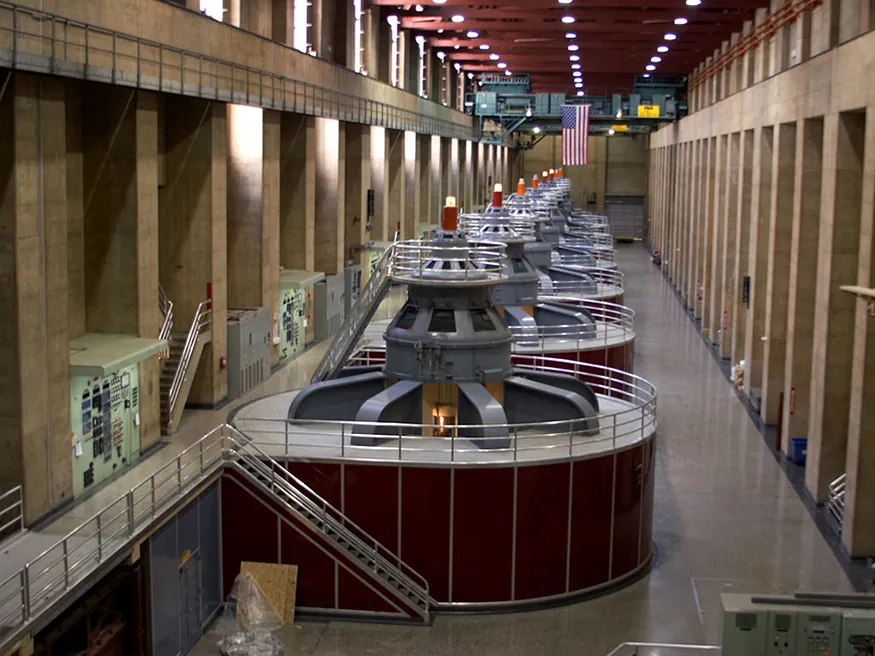 The interior of a large power generation facility, featuring multiple industrial-scale generators, an American flag, and robust concrete architecture.
