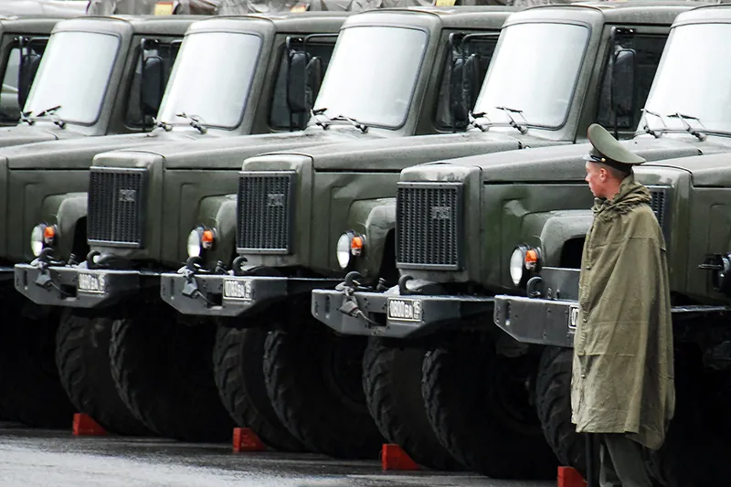 A person dressed in an olive green poncho and an olive green military hat stands in front of a row of 5 large military vehicles.
