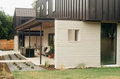 An image of the side and back of a house. The concrete on the side of the house shows lines from building layers of concrete in the additive manufacturing process. 