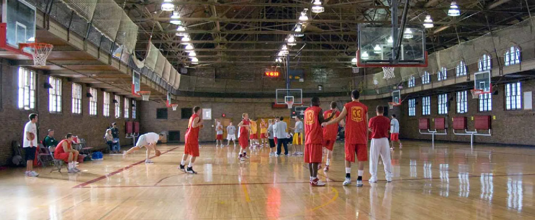 Basketball players in red and yellow uniforms practice in a spacious, classic gymnasium with high ceilings, brick walls, and multiple hoops. A scoreboard shows 16:33.