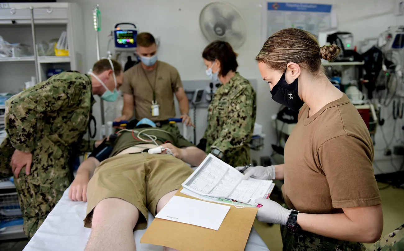 A photograph of military personnel examining patient in hospital bed.
