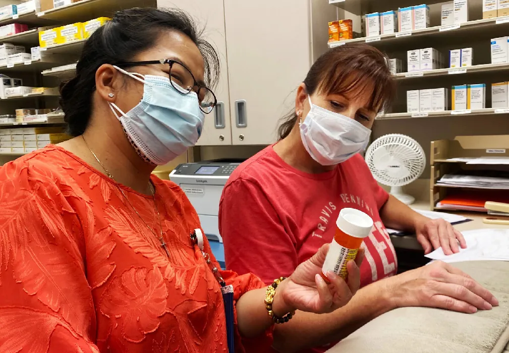 Two healthcare professionals sit next to each other looking at a prescription bottle that one of them hold up. The shelves behind them contain other boxes of prescription medications.