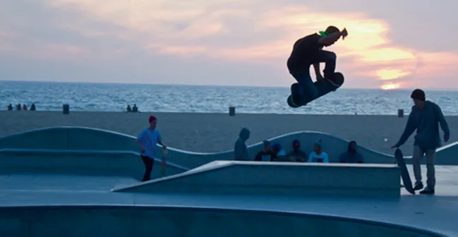 A skateboarder catches air in a concrete skatepark with the ocean and a sunset horizon in the background. Other people are seen relaxing on the beach and in the skatepark.
