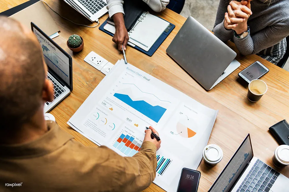 A group of people are at a table, with their hands shown holding pens. They're all pointing to data on a piece of paper.